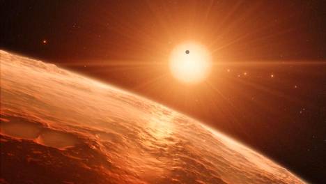 Taiteilijan näkemys vieraan aurinkokunnan reunamilta. Sieltä näkyy peräti seitsemän maapallon kaltaista planeettaa, eksoplaneettaa. Kaikki punertavat planeetat ja yksi musta kiertävät pientä kääpiötähteä, jonka nimi on Trappist-1. Piirroksen perustana on tähdestä ja planeetoista havaitut arvot.
