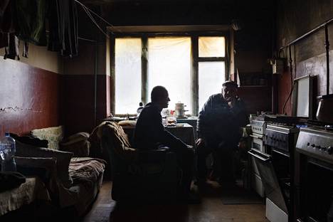 Дмитрий Янченко (слева) живёт в разрушенной многоэтажке в центре Изюма. Комната отапливается газовой плитой. Окно заделано пластиком, на стенах – копоть. В гостях у Дмитрий – друг Виктор. Фото: Саара Мансиккамяки / HS
