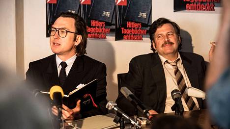 Toimittajat Gerd Heidemann (Lars Eidinger, vas) ja Felix Bloom (Hans Jochen Wagner) tyytyväisinä lehdistötilaisuudessa, jossa Hitler-skuuppia esitellään. 