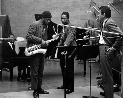 John Coltrane (kesk.) teki jazzin historiaa 9. joulukuuta 1964, mutta kukaan ei ollut tilannut paikalle valokuvaajaa. Seuraavana päivänä hän palasi samaan Rudy Van Gelderin studioon jatkamaan levyn A Love Supreme äänityksiä kokoonpanolla, johon kuului myös saksofonisti Archie Shepp. Vasemmalla Coltranen kvartetin vakiopianisti McCoy Tyner, oikealla levyn A Love Supreme tuottaja ja Impulse!-levy-yhtiön johtaja Bob Thiele.
