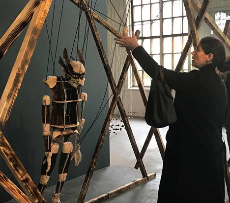 Hidden-näyttelyn kuratoinut Laura Sarvilinna näyttää, miten iso puumarionetti seuraa liikkeitä. Hidden Puppeteer -teoksen (2022) tekijä on Aapo Repo.