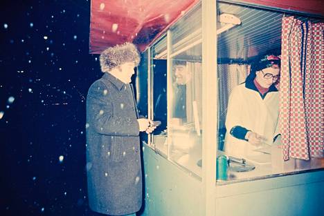 Mies odotti lumisateessa grilliruokaa nakkikioskilla 1970-luvulla.