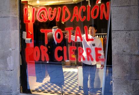 Barcelonassa asiakas sovittaa paitaa kaupassa, jonka ikkunaan on maalattu ”Täysi loppuunmyynti” maaliskuussa 2021. Monet liikkeet joutuivat lopettamaan toimintansa koronan vuoksi. 
