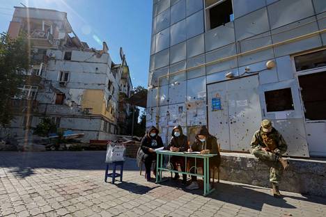 Vaalikomission jäsenet odottavat sotilaan kanssa ulkona tuhoutuneen asuinrakennuksen edessä Mariupolissa.
