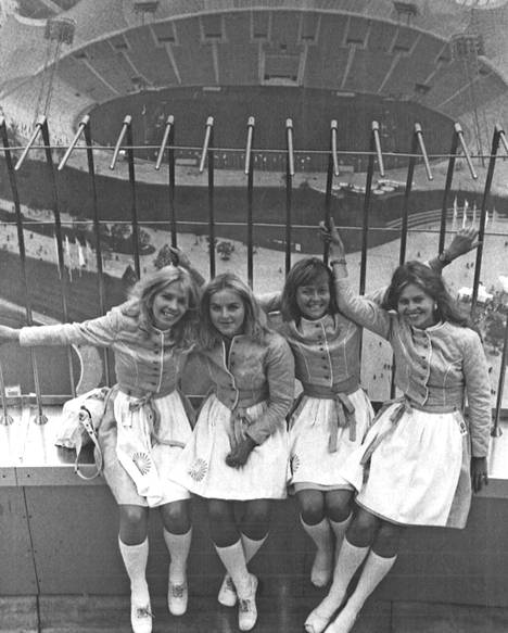 Suomea puhuvat kisaemännät Ritva-Maria Belz (vas.) Rita Ingberg, Sirpa Kukkonen ja Ulla Laakso ovat istahtaneet olympiatornin huipulle. Taustalla näkyy olympiastadion. Kaikki yli tuhat kisaemäntää ovat pukeutuneet yhtenäiseen sinivalkoiseen asuun.