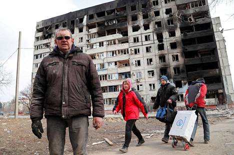 Yksihanskainen mies ja tavaroitaan kuljettavia ihmisiä kuvattuna tuhoutuneen kerrostalon edessä Mariupolissa torstaina.