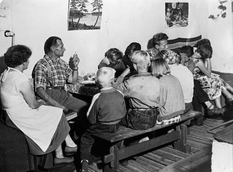 Osa 13 hengen perheestä aterioimassa pienen pöydän ääressä vuonna 1955 helsinkiläisessä parakkiasunnossa.