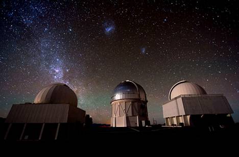 Kuu valaisee Cerro Tololon observatorion kupoleita Chilen autiomaassa. Yläpuolella kohoavat Suuri ja pieni Magellanin pilvi (keskellä) ja Linnunradan keskus (vasemmalla).