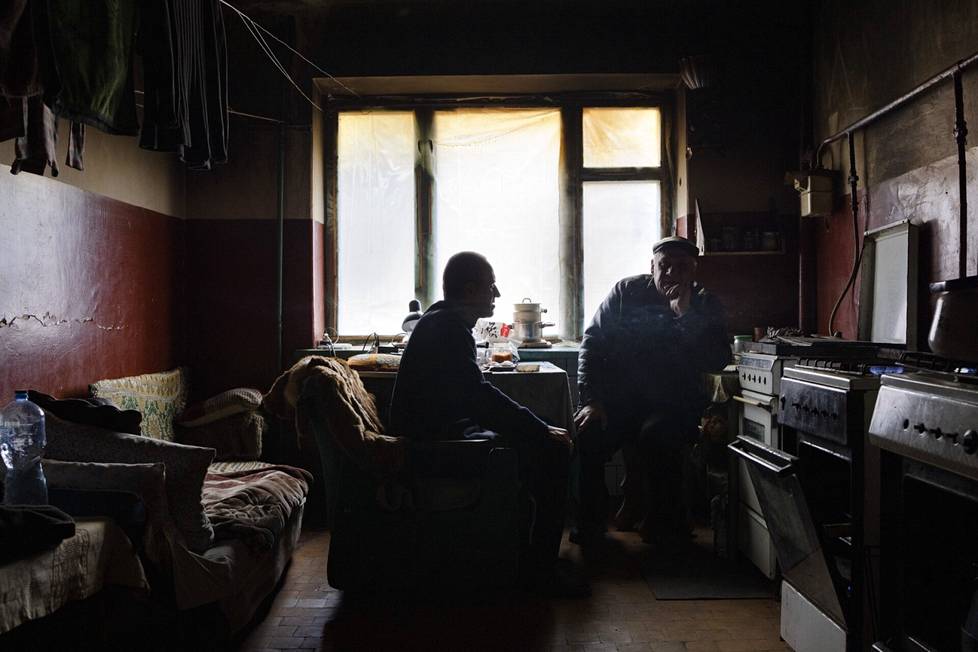 Dmytro Jantšenko (vas.) asuu pahasti tuhoutuneessa kerrostalossa Izjumin keskustassa.  Huonetta lämmitetään kaasulieden liekillä. Ikkuna-aukko on peitetty muovilla, ja seinät ovat noessa. Ystävä Viktor oli vierailulla.