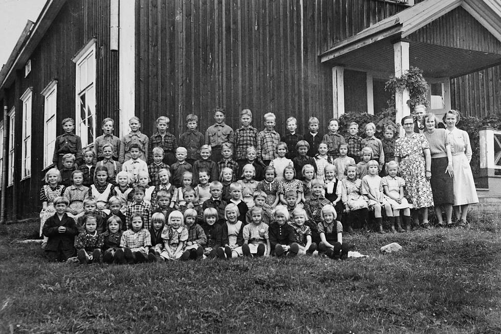 Ilomantsin Huhmarisvaaran koulun oppilaita ja henkilökuntaa kyläkoulun pihalla koulun päädyssä 1950-luvulla. Pirkko Kuivalainen istuu nurmella eturivissä neljäntenä vasemmalta.